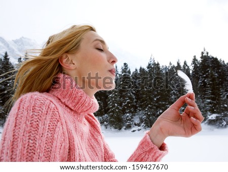 Close up profile portrait of an attractive young woman holding a light white feather in her hand and blowing on it while on a snow mountain vacation during a sunny winter day, outdoors.