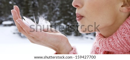 Close up profile portrait of an attractive young woman hand holding a light white feather and her lips blowing on it while on a snow mountain vacation during a cold winter day, outdoors.