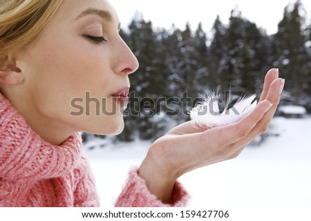 Close up profile portrait of an attractive young woman holding a light white feather in her hand and blowing on it while on a snow mountain vacation during a cold winter day, outdoors.