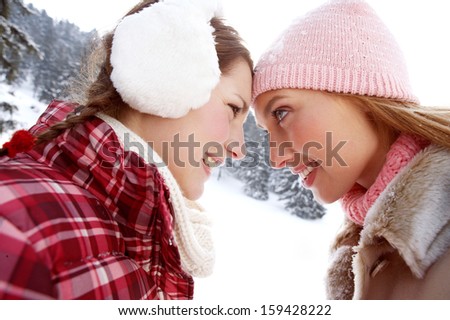 Two joyful young women friends enjoying a holiday in the snow mountains forest, smiling and holding their foreheads together, having fun during a cold winter day, outdoors.
