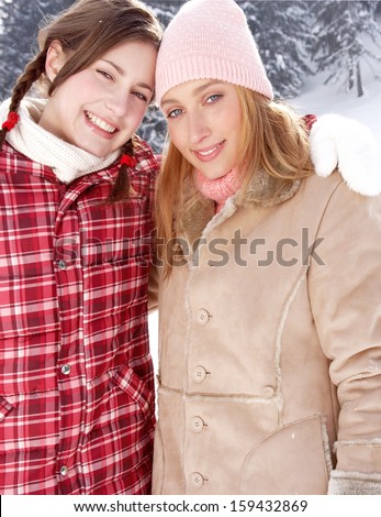 Two young friends girls with their arms around each others shoulders, joyfully smiling at the camera with their heads together and enjoying a sunny winter day in the snow mountains.