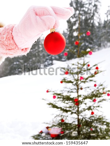 Close up detail view of a woman hand wearing a pink glove and holding a red decoration bar ball with care while setting up a christmas tree with gifts in the snow mountains wilderness.