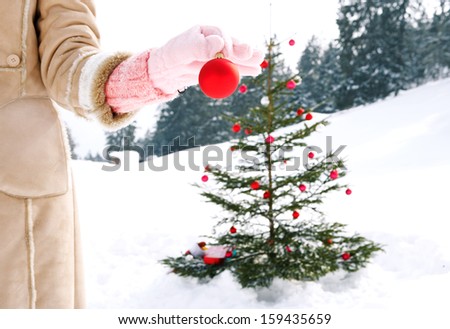 Close up detail view of a woman hand wearing a pink glove and holding a red decoration bar ball with care while setting up a christmas tree with gifts in the snow mountains wilderness.