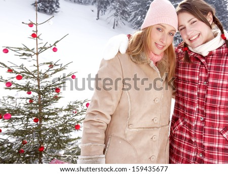 Two young friends girls with their arms around each others shoulders, joyfully smiling at the camera, celebrating christmas with a decorated xmas tree in the snow mountains.