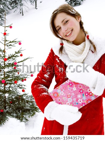 Attractive girl celebrating Xmas and holding a gift box while in the snow mountains with a natural decorated christmas tree with bar balls during a cold winter day, outdoors.