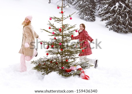 Two young joyful and attractive girls friends playing, chasing and running around a decorated christmas tree in the snow mountains nature, celebrating xmas and having fun together.