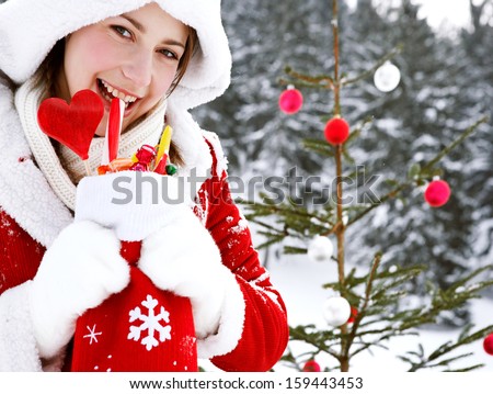 Close up portrait of an attractive young woman with a Christmas tree with bar balls, holding a sock full of candy sweets and biting one, smiling in the snow mountains outdoors.