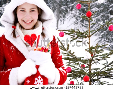 Close up portrait of an attractive young woman with a natural Christmas tree with bar balls, holding a sock full of candy sweets and joyfully smiling in the snow mountains outdoors.