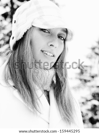 Black and white close up portrait of a beautiful young blond woman wearing fashionable clothes and smiling at the camera while visiting the snowed mountains and trees, outdoors with nature.