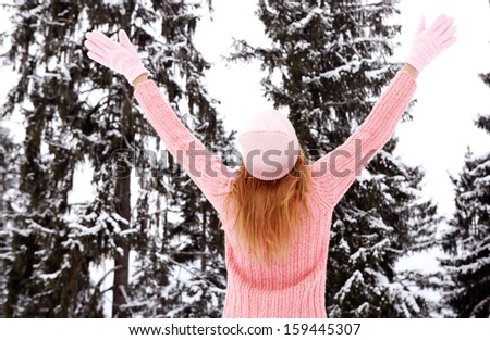 Rear view of a young woman with blond hair and a pink jumper, hat and gloves with her arms raised up in the air, enjoying the snow trees in the winter mountains, outdoors.