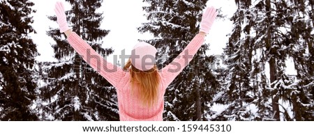 Horizontal rear view of a young woman with blond hair and a pink jumper, hat and gloves with her arms raised up in the air, enjoying the snow trees in the winter mountains, outdoors.