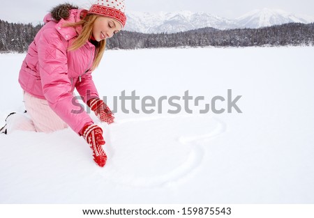 Attractive young woman kneeling on a frozen lake in the snow mountains forest landscape, drawing a heart on the snow with her hands during a skiing winter holiday, outdoors.