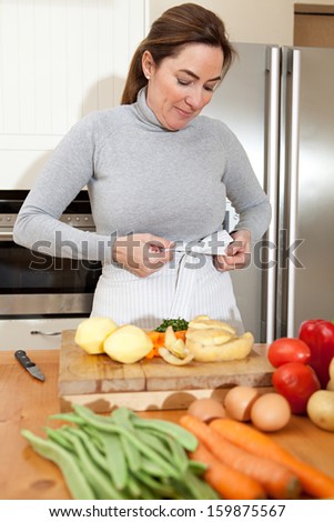 Attractive mature woman tying up her apron bow while peeling potatoes and cooking vegetables in the kitchen at home, with healthy organic produce and smiling, indoors.