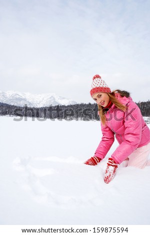 Attractive young woman kneeling on a frozen lake in the snow mountains forest landscape, drawing a heart on the snow with her hands during a skiing winter holiday, outdoors.