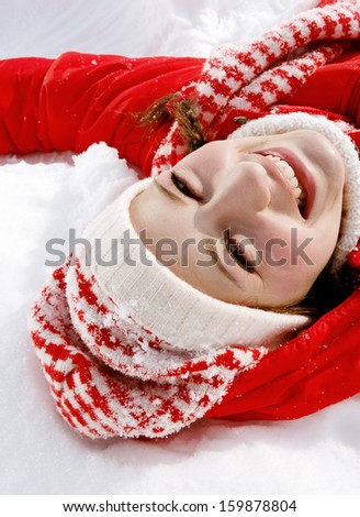 Over head view of a beautiful young woman laying down on the snow moving her arms up and down creating a snow angel figure, playing games while on a sunny winter vacation.