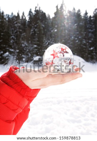 Part section of a woman hands holding a snow globe with red and white glitter stars floating inside, while in the snow mountains during a sunny winter vacation, outdoors.