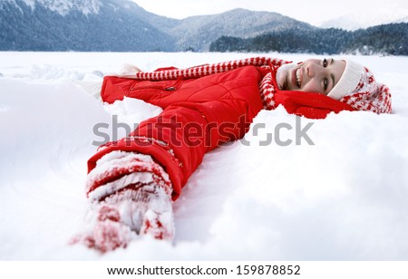 Side view of a beautiful young woman laying down on a frozen snow lake moving her arms up and down creating an angel figure shape, playing games during a sunny winter  vacation.