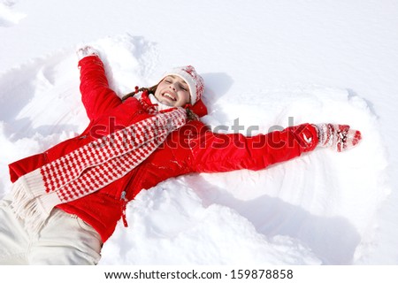 Beautiful joyful young woman laying down on a frozen snow lake moving her arms up and down creating an angel figure shape, playing games while on vacation during a sunny winter day.