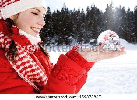 Side close up portrait of a joyful young woman holding a snow globe with red and white glitter stars floating inside, while in the snow mountains during a winter vacation, outdoors.