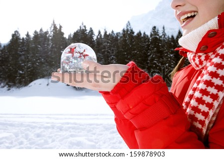 Side close up portrait of a joyful young woman holding a snow globe with red and white glitter stars floating inside, while in the snow mountains during a winter vacation, outdoors.
