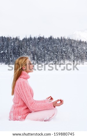 Side view of an attractive young woman sitting in a yoga position in a frozen lake in the snow mountains landscape, meditating and contemplating the scenery during a cold winter day outdoors.
