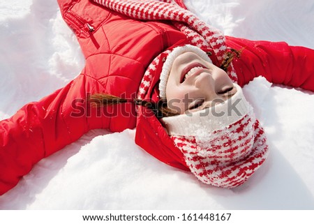 Over head view of a young woman laying down on a frozen snow lake moving her arms up and down creating an angel figure shape, playing games while on vacation during a sunny winter day.