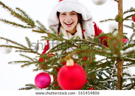 Portrait of an attractive young woman having fun and playing in the snowed mountains, decorating a small Christmas tree with bar balls, while joyfully smiling in the snow, outdoors.