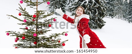 Panoramic side view of an attractive girl celebrating Christmas and decorating a natural Xmas tree with bar balls while on holiday in the skiing snow mountains during a cold winter day outdoors.