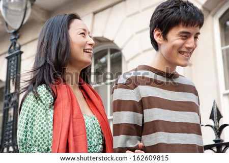 Close up portrait of a Japanese young couple enjoying a holiday in the city of London, sightseeing and walking passed a railing in a classic stone buildings street, laughing with fun outdoors.