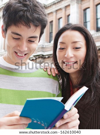 Close up portrait of a young and attractive Japanese tourist couple visiting the city of London on holiday together and reading a guide book in a classic street, smiling joyfully outdoors.
