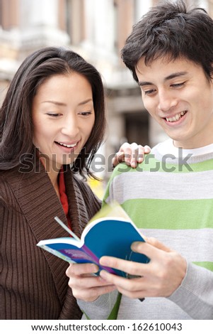 Close up portrait of a young and attractive Japanese tourist couple visiting the city of London together on holiday and reading a guide book in a classic street, smiling joyfully outdoors.
