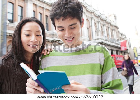 Portrait of a young and attractive Japanese tourist couple visiting the city of London together and reading a guide book in a classic street, smiling joyfully outdoors.