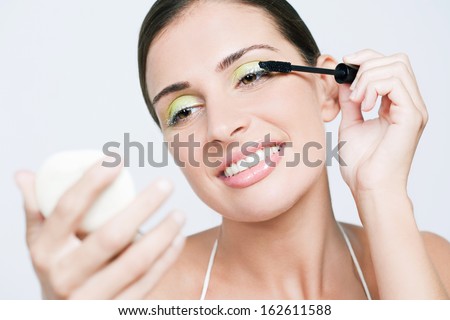 Close up portrait of a beautiful young woman applying party make up and glitter mascara on her eyelashes using a hand mirror and smiling, isolated against a white background, indoors.