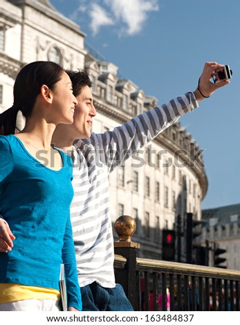 Side view of an attractive Japanese tourist couple using a consumer digital photo camera to take pictures of themselves while visiting a London landmark street during a sunny day, outdoors.
