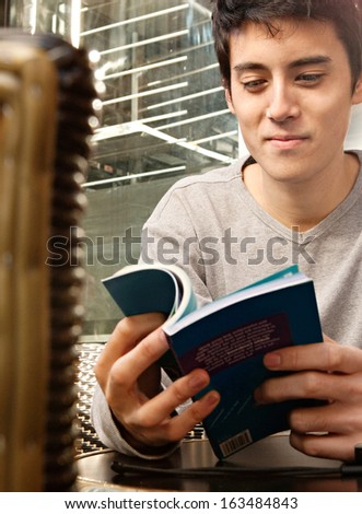 Attractive young Japanese tourist man sitting at a coffee shop in the city of London, having a coffee and reading a travel guide book, smiling outdoors during a sunny day.