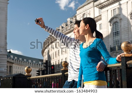 Side View Of An Attractive Japanese Tourist Couple Using A Consumer Digital Photo Camera To Take Pictures Of Themselves While Visiting A London Landmark Street During A Sunny Day, Outdoors.