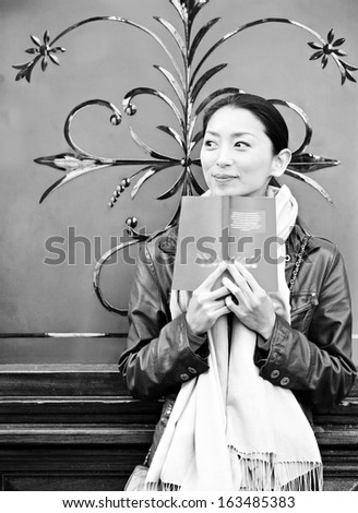 Black and white portrait of an attractive Japanese tourist woman in the city of London, reading a travel guide book, leaning on a decorative glass and wood frame in an English pub building.