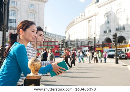 Side portrait view of a young Japanese tourist couple in Piccadilly Circus landmark street with red buses, while visiting the city of London on holiday, smiling during a sunny day outdoors.