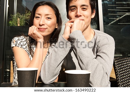 Happy Japanese couple sitting together at a coffee shop terrace having a hot coffee beverage and leaning on their hands while smiling at the camera during a sunny day, outdoors.