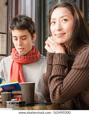 Portrait of a young Japanese couple sitting down at a cafe shop terrace taking a break in the city of London while on vacation, drinking a cup of hot coffee and reading a travel guide book, outdoors.