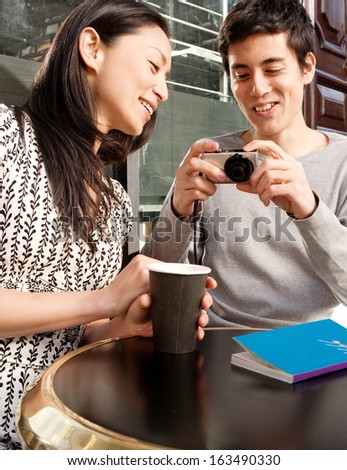 Joyful Japanese tourist couple sitting at a cafe terrace shop in London city, using a consumer digital photo camera to look at their trip pictures, smiling outdoors during a sunny day.