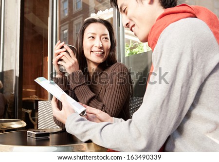Young and attractive Japanese couple sitting down at a cafe shop terrace in  the city of London while on vacation, drinking a cup of hot coffee and reading a travel guide book, smiling outdoors.
