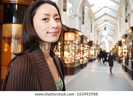 Portrait of an attractive Japanese tourist woman smiling and visiting a shopping mall in the city of London while on a holiday break trip, with stores and lights in the background.