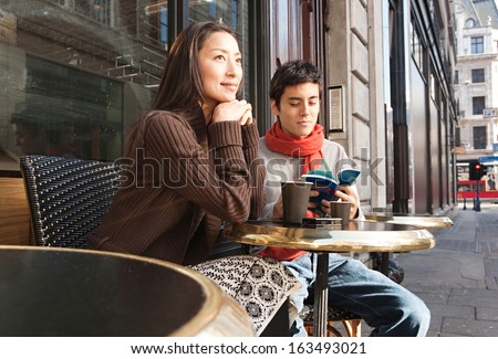 Attractive Japanese couple sitting down at a cafe shop terrace taking a break in  the city of London while on vacation, drinking a cup of hot coffee and reading a travel guide book, outdoors.