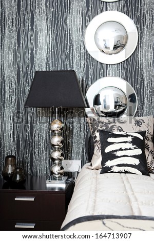 Designer interior detail view of a luxury hotel bedroom with elegant decorative mirrors and bedside lamp, rich textures and fabrics and organic graphic wallpaper. Home interior with no people.