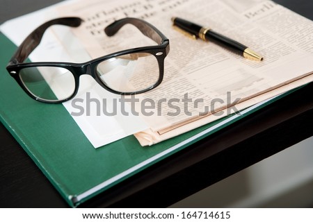 Close up still life of a pair of reading glasses and a black pen laying on an orange financial newspaper and notebook on a dark wooden writing desk. Office interior with no people.