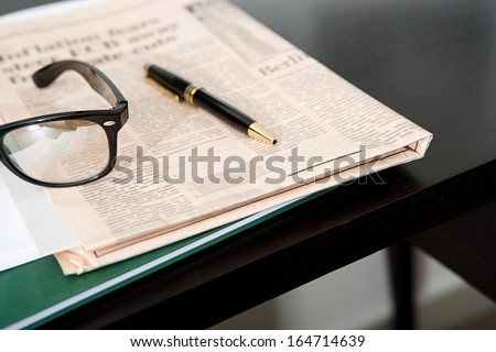 Close up still life of a pair of reading glasses and a black pen laying on an orange financial newspaper and notebook on a dark wooden writing desk. Office interior with no people.