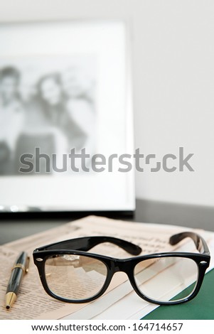 Close up still life of a pair of reading glasses and a black pen laying on an orange financial newspaper on a dark wooden writing desk with a family photo frame. Office interior with no people.