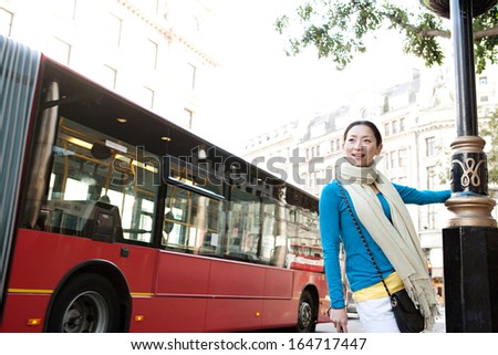 Young and attractive Japanese tourist woman enjoying sightseeing in the destination city of London with red buses and classic buildings around her, during a sunny day on holiday, outdoors.