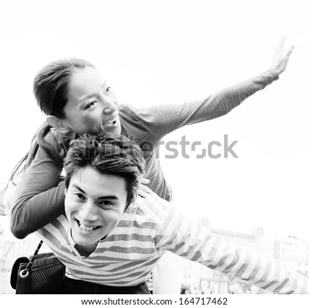 Black and white portrait of a joyful Japanese couple being playful with the man carrying the woman in a piggy bag, laughing with their arms open while visiting London on holiday, outdoors.
